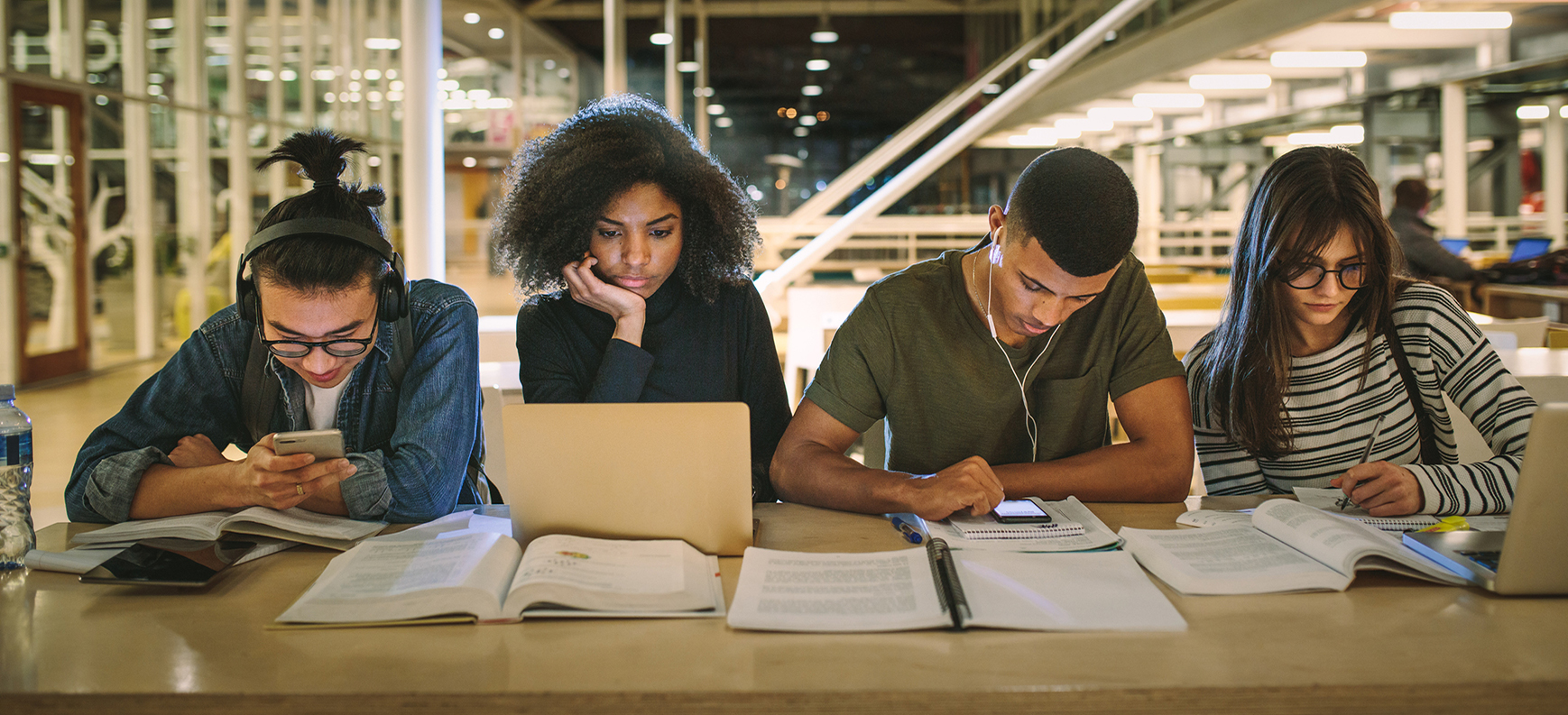 Four students studying using books, computers, pen and paper, and mobile devices.